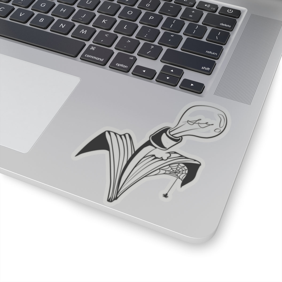 This is the Transparent 4" x 4" Storm Garden Studio Sticker. The sticker is a lightbulb screwed onto a plant with two leaves. The right leaf is bitten through with a spiderweb and a spider. The sticker is on a silver laptop and on the right of it. The picture shows the corner of it, to the left is the mousepad and the black keyboard on top. The laptop with the sticker is against a white blackground.