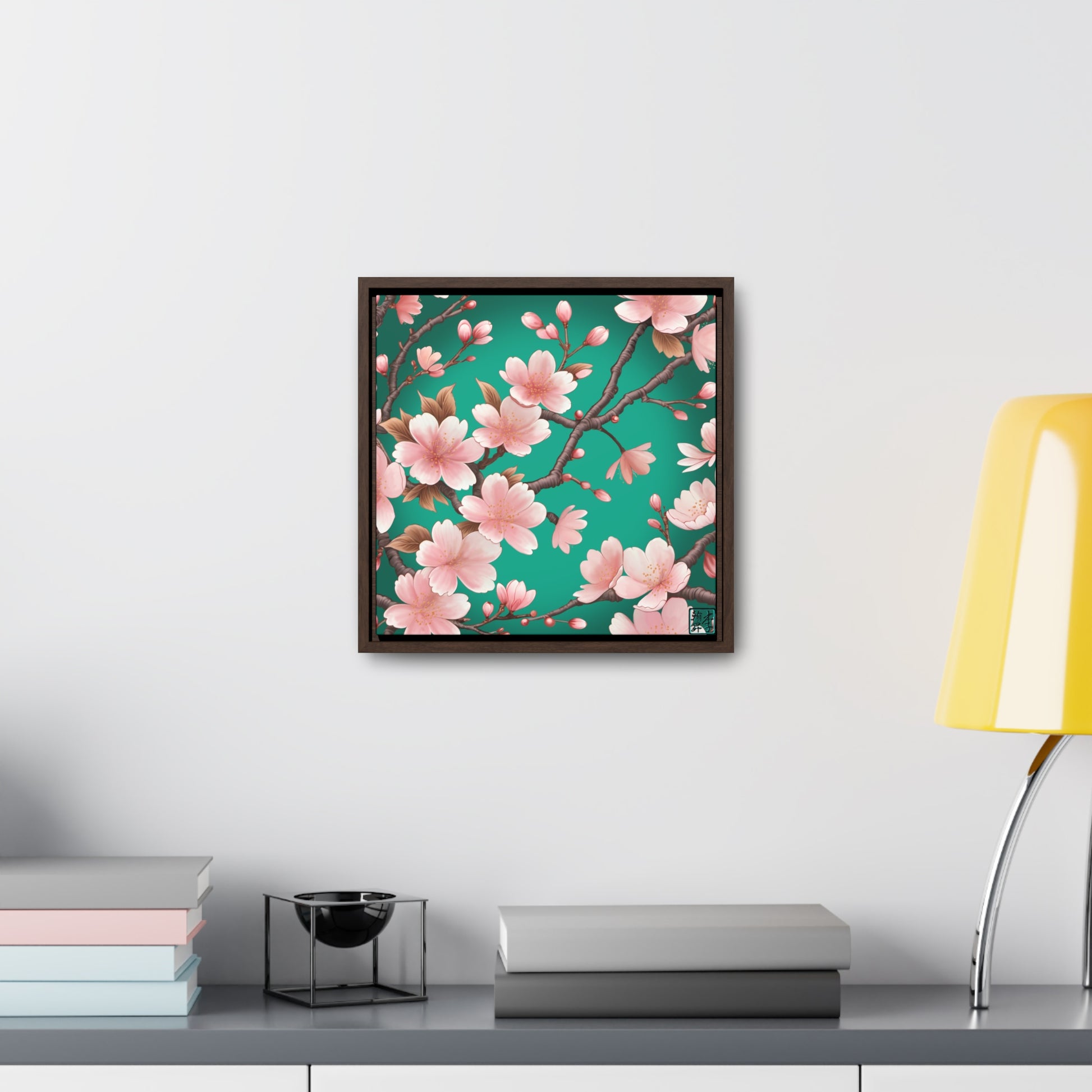 This is the Walnut 16" x 16" Sakura Gallery Canvas Wrap by Lee Hansheng Studios. The illustration has a blue/teal background, 3-4 thin dark wood branches with pink/light pink buds on each stem. There are blooming and already flowered sakuras on each branch. The studio's logo is on the bottom right printed in black outlined in blue. Below the canvas is a gray/white shelf, yellow lamp, a decoration, gray and pastel books on the shelf spread across it. The canvas is on a white wall.