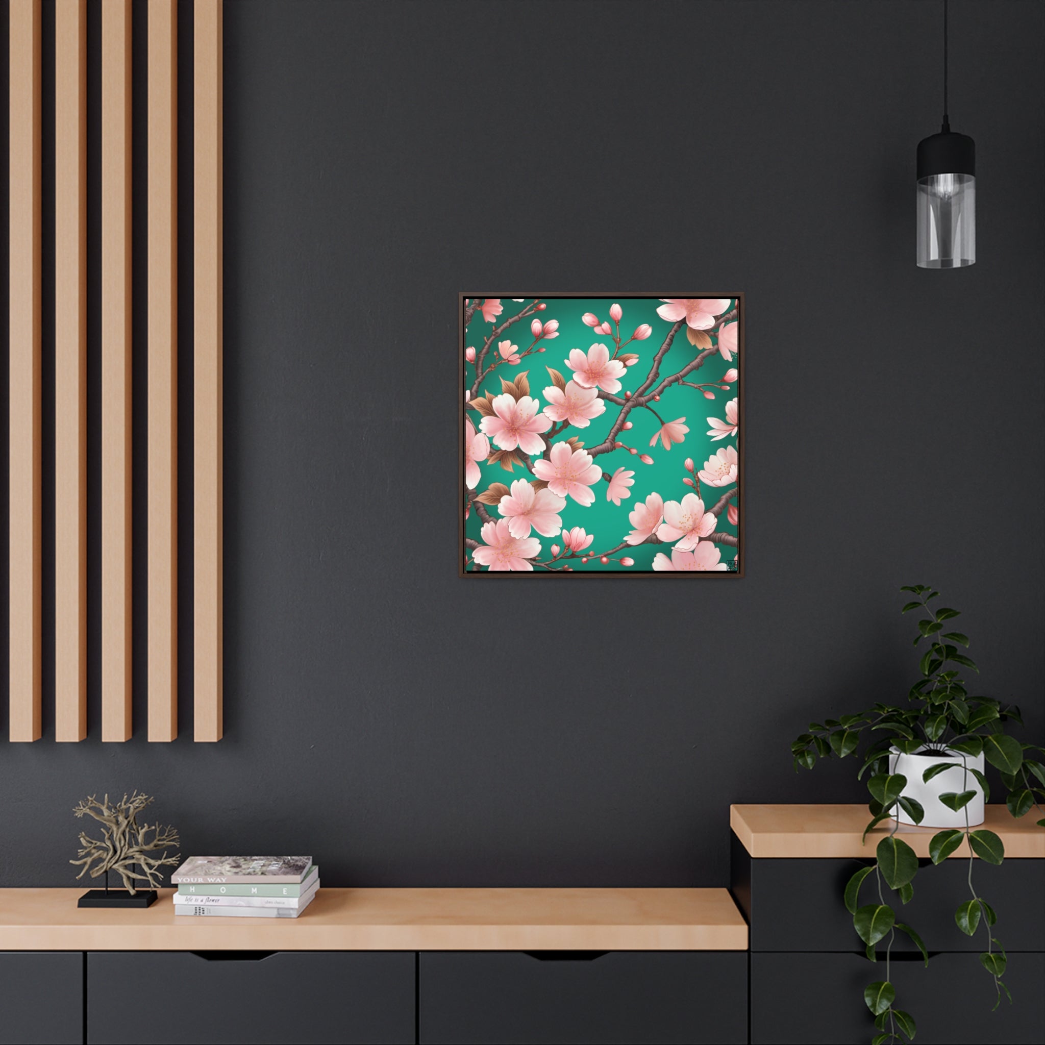 This is the Walnut 12" x 12" Sakura Gallery Canvas Wrap by Lee Hansheng Studios. The illustration has a blue/teal background, 3-4 thin dark wood branches with pink/light pink buds on each stem. There are blooming and already flowered sakuras on each branch. The canvas is against a black wall, wood panels to the left, hanging black lamp to the right, black and wood shelves below. There are assorted decorations on each side like books, tree decor and an ivy plant with a white pot. 