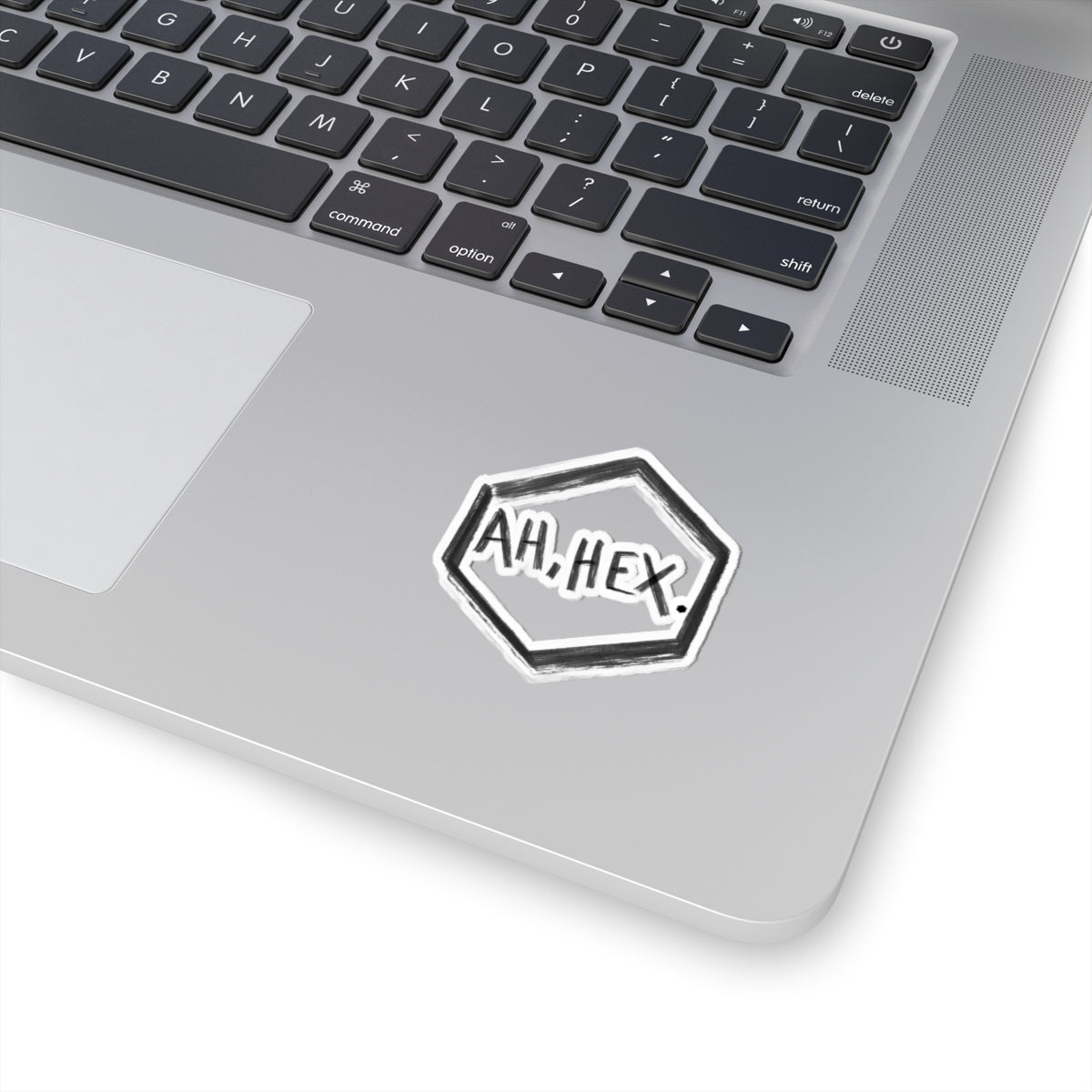 This is the White 2" x 2" Ah, Hex Kiss-Cut Sticker by Tidy Tea Witch. The writing is in black and says. "AH, HEX." in the middle of the sticker. There is no illustration or image on it but is hexsagon shaped. The sticker is on a gray laptop with the black and white keyboard above and a gray/white mousepad to the left. The image is against a white background.