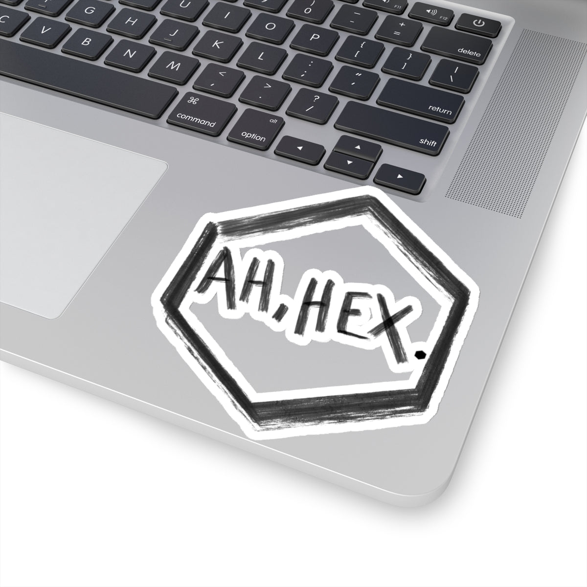 This is the White 4" x 4" Ah, Hex Kiss-Cut Sticker by Tidy Tea Witch. The writing is in black and says. "AH, HEX." in the middle of the sticker. There is no illustration or image on it but is hexsagon shaped. The sticker is on a gray laptop with the black and white keyboard above and a gray/white mousepad to the left. The image is against a white background.