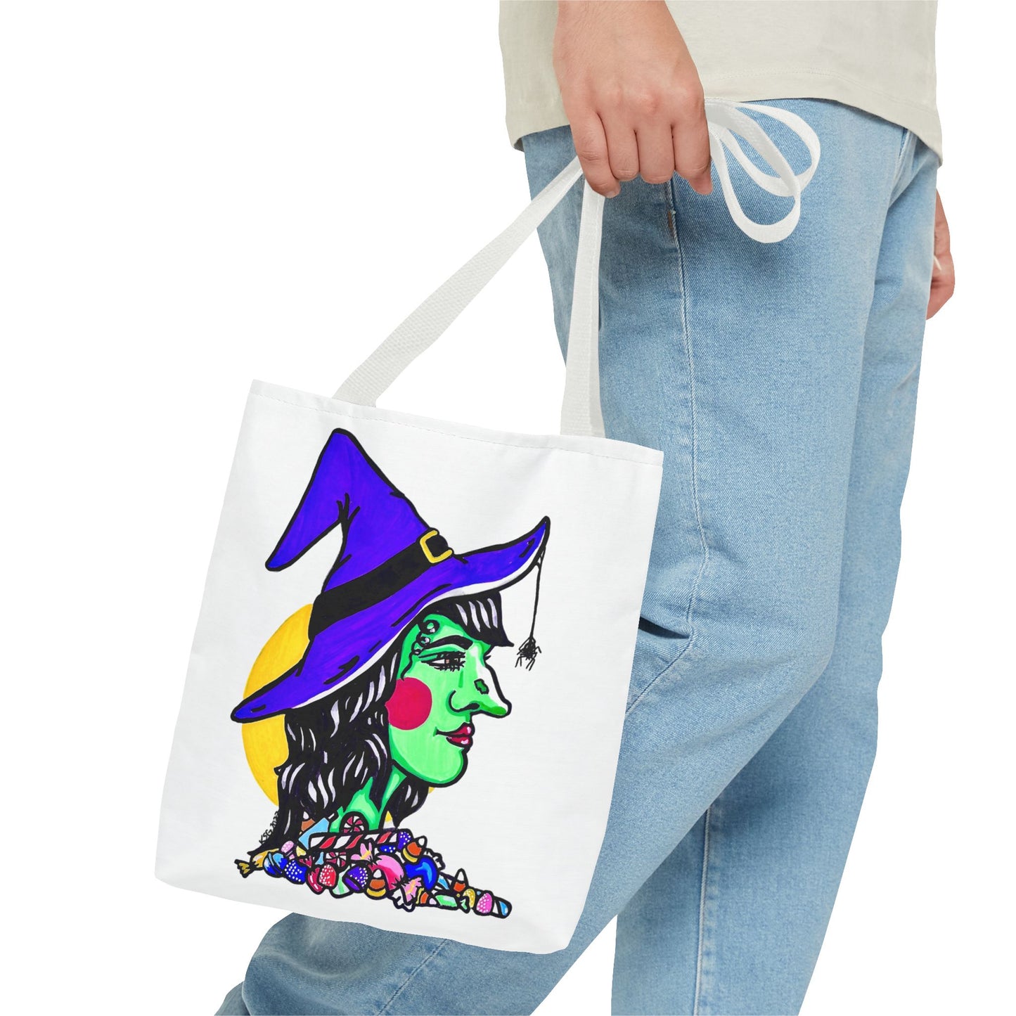 This is the White 13" x 13" Witch Tote Bag by Storm Garden Studio. The handles are color matched. It shows the side profile of a witch. She has a purple witch hat, a black belt on hat, yellow buckle, a spider crawling down the front, red rosy cheeks, red lips and assorted candy under the neck. Behind her is a bright yellow circle past her hair. A man's hand with a beige tee and light- colored jeans is grabbing the handles from the hip down. The tote is against a white background.