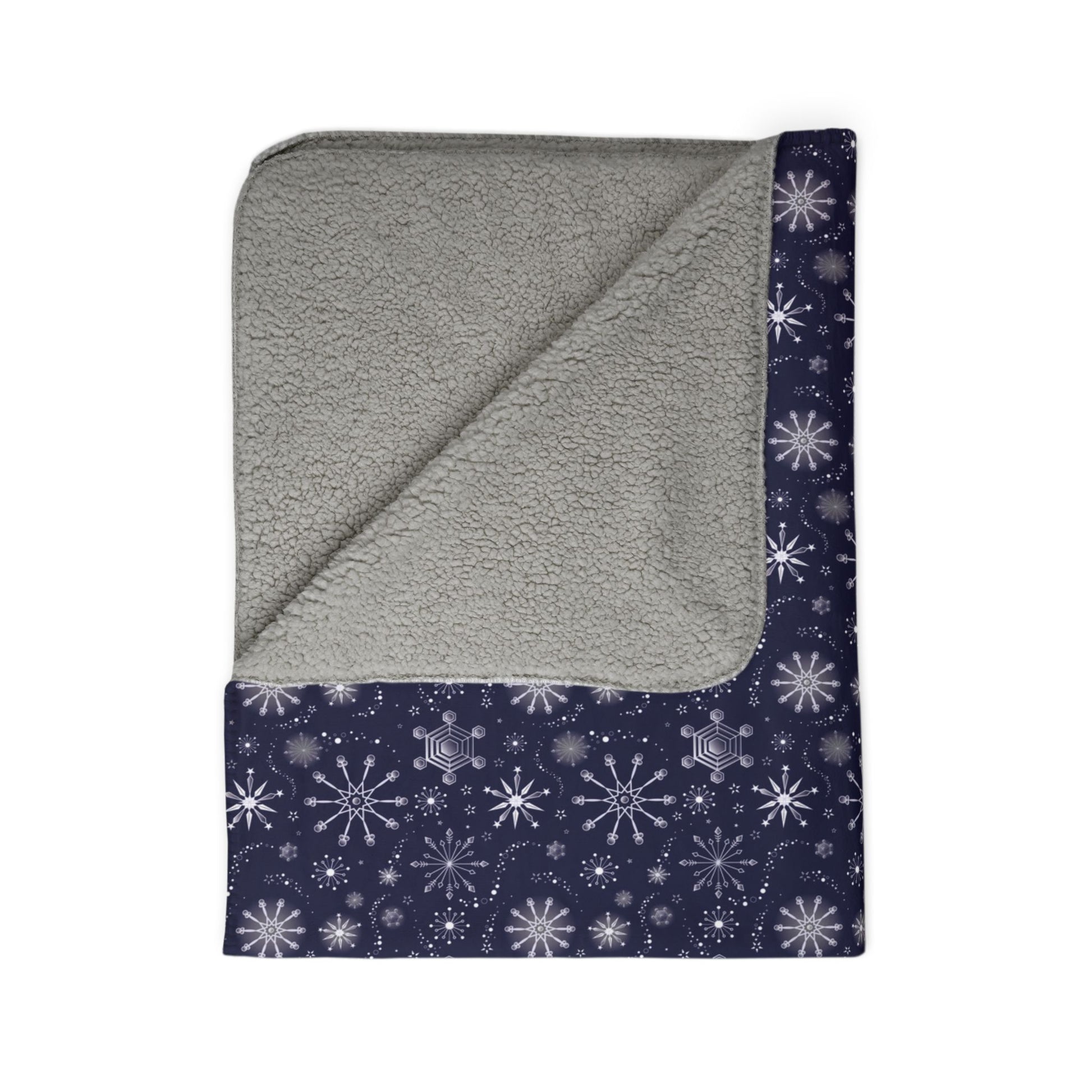 This is the grey 60" x 80" Winter Blizzard Sherpa Blanket. The blanket is folded. It is then revealed at the corner to show the inside sherpa and color. The background is white. The design are different types of snowflakes with different sizes of circles within each flake. The blanket is dark blue and the snowflakes and design are all white and spread throughout.