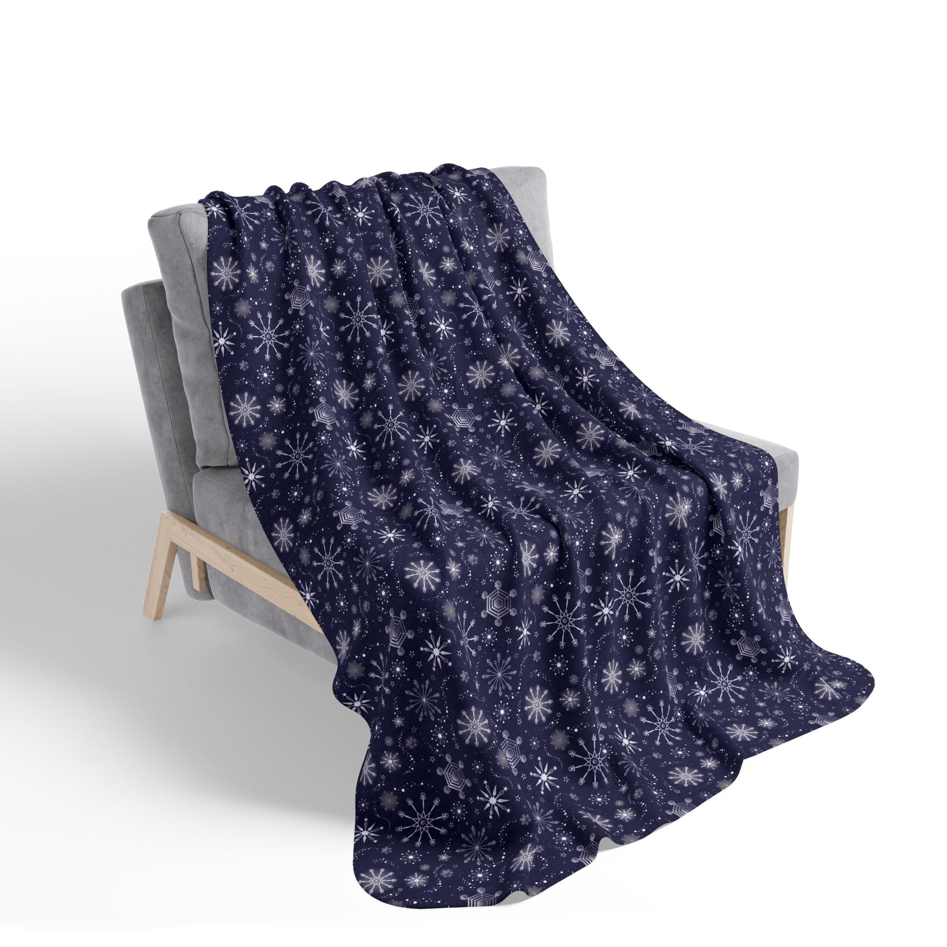 This is the beige 60" x 80" Winter Blizzard Sherpa Blanket. The blanket is placed on a grey fabric lounge chair with light wood legs. The background is white. The design are different types of snowflakes with different sizes of circles within each flake. The blanket is dark blue and the snowflakes and design are all white and spread throughout.